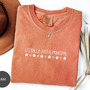 May include: A coral-colored t-shirt with the text "LITERALLY JUST A PRINCIPAL" in white, with small flower graphics. The shirt is folded, and a gold necklace is draped across it. A brown hat is visible in the background.