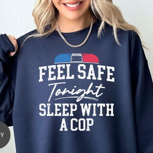 May include: Navy blue sweatshirt with the text "FEEL SAFE Tonight SLEEP WITH A COP" in white. The design includes a red and blue police light graphic. The sweatshirt is a crew neck style.