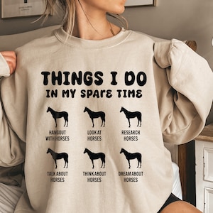 May include: A beige sweatshirt with the text "THINGS I DO IN MY SPARE TIME" and six black silhouettes of horses with text describing activities related to horses. The activities are: Hangout with horses, Look at horses, Research horses, Talk about horses, Think about horses, Dream about horses.