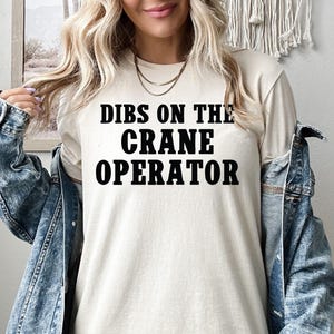 May include: A white t-shirt with the text "DIBS ON THE CRANE OPERATOR" printed in black.