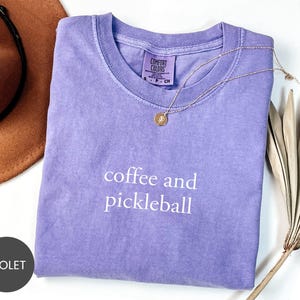 May include: A violet-colored Comfort Colors t-shirt with the words "coffee and pickleball" in white. A gold necklace with a pendant rests on the shirt. A brown hat and dried palm leaves are also in the image.