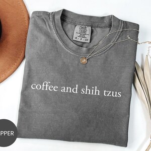 May include: A gray Comfort Colors t-shirt with the text "coffee and shih tzus" in white. A gold necklace with a pendant is draped across the shirt. A brown hat and decorative leaves are in the background.
