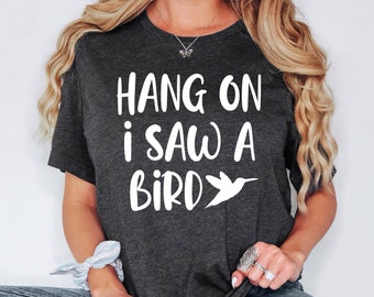 May include: A dark gray t-shirt with the text "HANG ON i SAW A BIRD" in white lettering. A white silhouette of a hummingbird is next to the text.