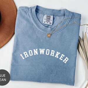 May include: A blue jean colored t-shirt with the word "IRONWORKER" in white block letters. A gold necklace with a pendant is draped across the shirt. The shirt is folded and placed on a white surface.