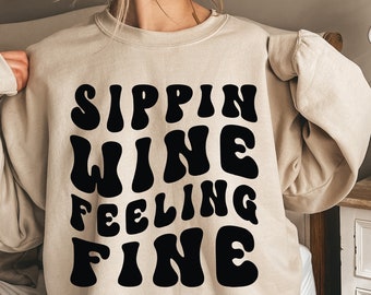 Pode incluir: Uma camisa de moletom bege com o texto "Sippin Wine Feeling Fine" em letras pretas de estilo retrô.