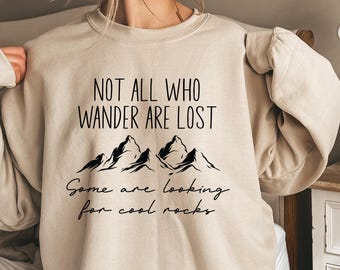 May include: A beige sweatshirt with a black graphic print that reads "Not all who wander are lost. Some are looking for cool rocks." The graphic also includes a stylized image of mountains.