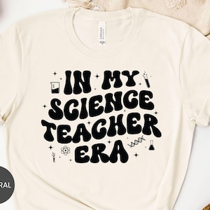 May include: Cream-colored t-shirt with the text "IN MY SCIENCE TEACHER ERA" in bold, black, retro-style lettering. The design includes science-themed icons like beakers, test tubes, and a DNA strand. The shirt is made of a soft material.