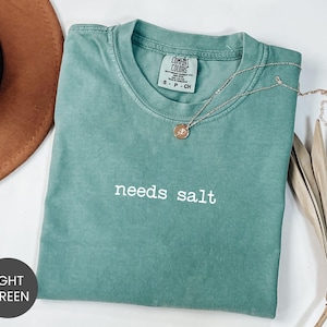 May include: Light green t-shirt with the phrase "needs salt" in white lettering. The shirt has a crew neck and short sleeves. A silver necklace with a small pendant is draped across the shirt.