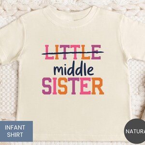 May include: A natural-colored infant t-shirt featuring the text LITTLE with a line through it, followed by the word middle in a script font, and the word SISTER in block letters. The text uses a color palette of pink, orange, and dark blue.