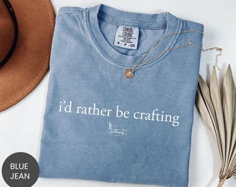 Puede incluir: Una camiseta azul vaquera Comfort Colors con la frase "i'd rather be crafting" en blanco. Un pequeño gráfico de un sofá está debajo del texto. Un collar dorado con un colgante está drapeado sobre la camisa.