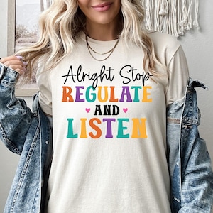 May include: Cream-colored t-shirt with the words "Alright Stop REGULATE and LISTEN" in colorful block letters. The shirt is worn with a denim jacket.
