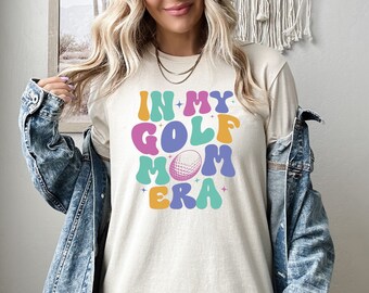 May include: A beige t-shirt with the text "In My Golf Mom Era" in colorful, retro-style letters. A golf ball is featured in the center of the design.