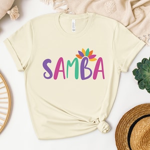 May include: A cream colored t-shirt with the word "SAMBA" in colorful letters and a multi-colored flower design above the word.