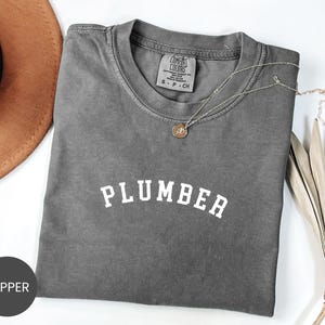 May include: A gray Comfort Colors t-shirt with the word "PLUMBER" in white block letters. A gold necklace with a pendant rests on the shirt. A brown hat is visible in the upper left corner. The shirt is a size S-P-CH.