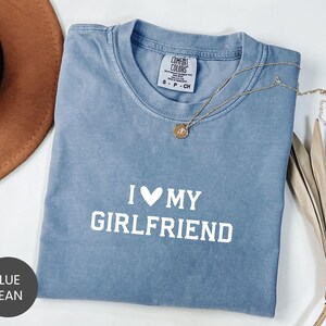 May include: Blue jean colored t-shirt with the text "I ♥ MY GIRLFRIEND" in white. A gold necklace with a pendant is draped across the shirt. The shirt is folded and has a tag that says "Comfort Colors".