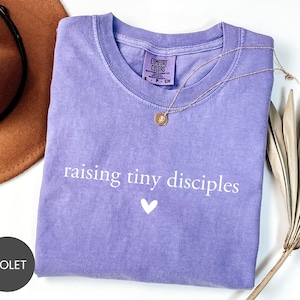 May include: A violet Comfort Colors t-shirt with the phrase "raising tiny disciples" and a white heart graphic. A gold necklace with a pendant rests on the shirt. A brown hat and dried plant are in the background.