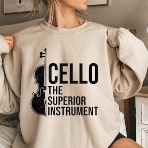 May include: Beige sweatshirt with a black cello graphic and the text "CELLO THE SUPERIOR INSTRUMENT". The design features a stylized cello silhouette. The sweatshirt has a crew neck and long sleeves.