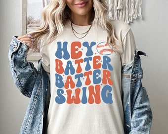 May include: A white t-shirt with a retro-style graphic that says "Hey Batter Batter Swing" in blue and orange letters. A baseball is featured above the text.