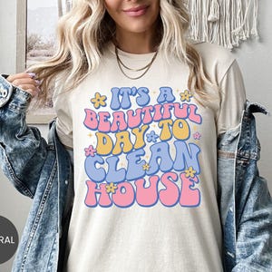 May include: A cream-colored t-shirt with the phrase "It's a beautiful day to clean house" in a retro font. The text is in shades of pink, blue, and yellow, with flower accents. The shirt is a casual style.
