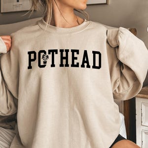 May include: A beige sweatshirt with the text "POTHEAD" printed in black. The word "POT" has a small plant icon in the "O".