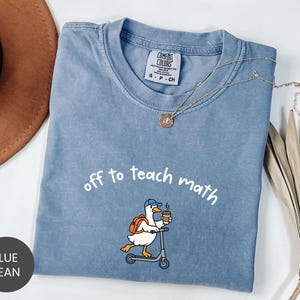 May include: A blue jean colored t-shirt featuring a graphic of a white duck wearing a blue cap and an orange backpack, riding a scooter while holding a coffee cup. The text above the duck reads off to teach math in white lettering.