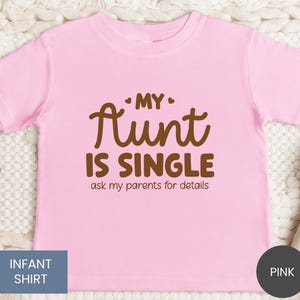 May include: A pink infant shirt with the text "MY AUNT IS SINGLE ask my parents for details" in brown lettering. The shirt is a solid pink color and has short sleeves. The words "INFANT SHIRT" are in a blue box.
