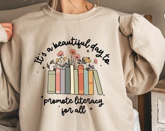 May include: A beige sweatshirt with a graphic print of a stack of colorful books with flowers on top. The text reads "It's a beautiful day to promote literacy for all."