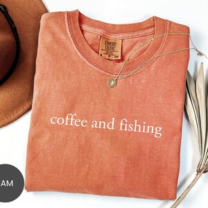 May include: A coral-colored t-shirt with the words "coffee and fishing" printed in white. A gold necklace and a brown hat are next to the shirt. The shirt is folded on a white surface.