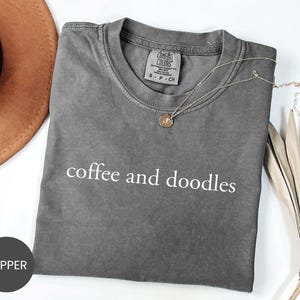 May include: A gray t-shirt with the words "coffee and doodles" printed in white. A gold necklace with a pendant is draped across the shirt. A brown hat and dried plant are in the background.