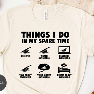 May include: Cream-colored t-shirt with the text "THINGS I DO IN MY SPARE TIME" in bold black letters. Below the text are six black icons depicting swimming-related activities, including swimming, watching, researching, talking, thinking, and dreaming about swimming.