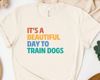 Puede incluir: Camiseta color crema con el texto "IT'S A BEAUTIFUL DAY TO TRAIN DOGS" en letras de colores. La camiseta está hecha de un material suave y tiene un diseño clásico de cuello redondo.