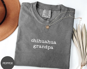 Op de afbeelding: Een grijs Comfort Colors t-shirt met de woorden "chihuahua grandpa" in het wit. Een gouden ketting met een hanger is over het shirt gedrapeerd. Een bruine hoed en een gedroogde plant staan op de achtergrond.