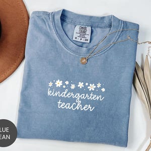May include: Blue jean colored t-shirt with the words "kindergarten teacher" in white script, adorned with small white daisy graphics. A gold necklace with a pendant rests on the shirt. The shirt is folded and displayed.