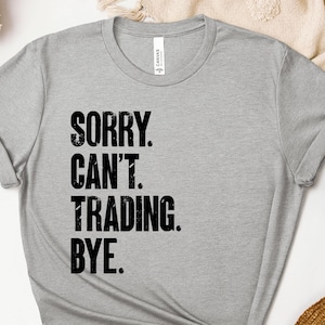 May include: A light gray t-shirt with the text "SORRY. CAN'T. TRADING. BYE." printed in black. The shirt has a crew neck and short sleeves. The shirt is made of a soft material.