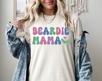 Könnte beinhalten: Ein hellbeiges T-Shirt mit dem Text "BEARDIE MAMA" in bunten, Retro-Lettern. Rechts neben dem Text befindet sich ein kleines grünes Eidechsen mit einem rosa Herzen.