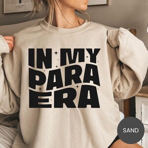 May include: Sand-colored sweatshirt with the text "IN MY PARA ERA" in bold black letters. The text is surrounded by small star shapes. The sweatshirt has a crew neck and long sleeves.
