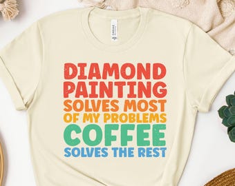 May include: Cream-colored t-shirt with the text "DIAMOND PAINTING SOLVES MOST OF MY PROBLEMS COFFEE SOLVES THE REST" in red, orange, green, and blue block letters. The shirt is made of a soft material.
