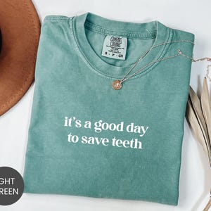 May include: A light green short-sleeve t-shirt with a crew neckline featuring the white printed text 'it's a good day to save teeth' on the chest. The garment includes a Comfort Colors brand label at the inner neck and a delicate gold-tone necklace.