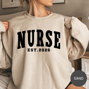May include: Sand-colored sweatshirt with the word "NURSE" in bold black letters across the chest, with "EST. 2026" below. The sweatshirt has a crew neck and long sleeves. The word "SAND" is in a circle in the bottom right corner.