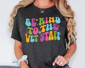 May include: Dark gray t-shirt with the text "BE KIND TO THE VET STAFF" in colorful, retro-style lettering. The design includes a paw print and flower accents. A casual, comfortable tee for veterinary staff supporters.
