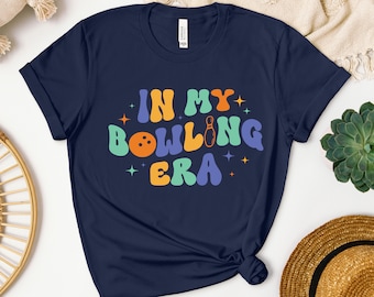May include: Navy blue t-shirt with the text "In My Bowling Era" in colorful retro-style letters. A bowling pin and ball are included in the design.