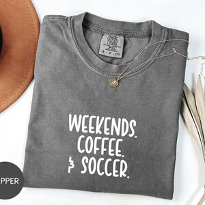May include: A charcoal gray short-sleeve t-shirt with the white text "WEEKENDS. COFFEE. & SOCCER." printed on the front. The shirt features a crew neckline and a Comfort Colors brand label on the inside collar. A small gold-toned pendant necklace rests on the fabric.