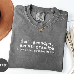 May include: A gray Comfort Colors t-shirt with the text "dad, grandpa, great-grandpa, I just keep getting better" in white. A gold necklace with a pendant is draped on the shirt. A brown hat is visible in the background.