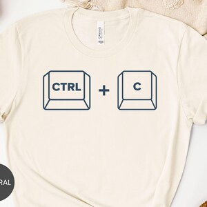 May include: A cream-colored short-sleeve t-shirt featuring a dark blue graphic print of two computer keyboard keys. The left key displays the text CTRL and the right key displays the letter C, separated by a plus sign in the center.