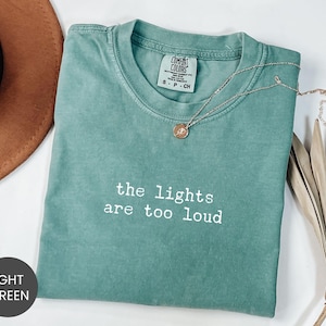 May include: A light green short-sleeve t-shirt with a crew neckline featuring the white embroidered text "the lights are too loud" on the chest. The garment has a Comfort Colors brand label on the inside collar indicating a size small.