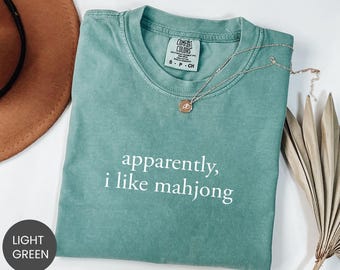 May include: Light green t-shirt with the text "apparently, i like mahjong" in white. The shirt has a crew neck and short sleeves. A gold necklace with a pendant rests on the shirt.