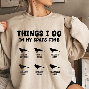 May include: A beige sweatshirt with the text "THINGS I DO IN MY SPARE TIME" and a series of black silhouettes of birds with text describing activities related to birds, such as "Hangout with Birds", "Look at Birds", "Research Birds", "Talk About Birds", "Think About Birds", and "Dream About Birds".