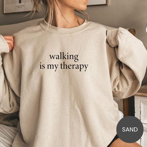 May include: A sand-colored sweatshirt with the phrase "walking is my therapy" printed in black. The sweatshirt has a crew neck and long sleeves. The image also includes a circular color swatch labeled "SAND".