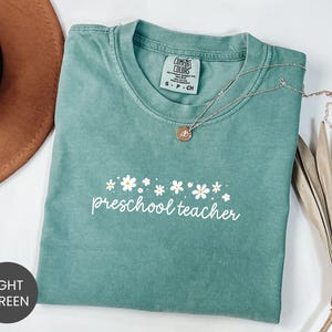 May include: A light green t-shirt with the words "preschool teacher" in white script, adorned with a daisy design. The shirt has a crew neck and short sleeves. A silver necklace with a pendant is draped across the shirt.
