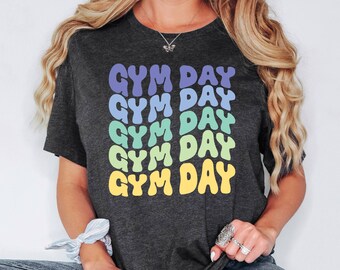 May include: Dark gray t-shirt with the text "GYM DAY" in a repeating pattern of colorful bubble letters. The text is in shades of purple, blue, green, and yellow. The shirt is a casual style.